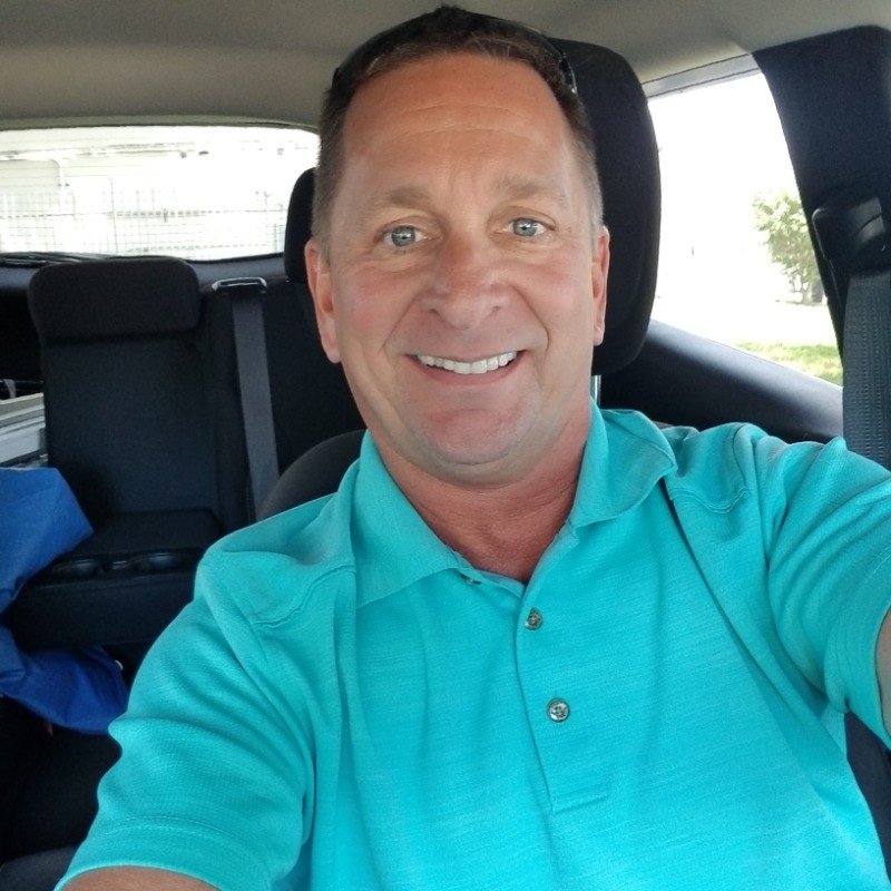 A smiling man wearing a teal polo shirt is sitting in the driver's seat of a car. The interior of the car is visible, with the back seat and some items in the back, including a blue bag. Sunlight is coming through the windows, illuminating the man's face.