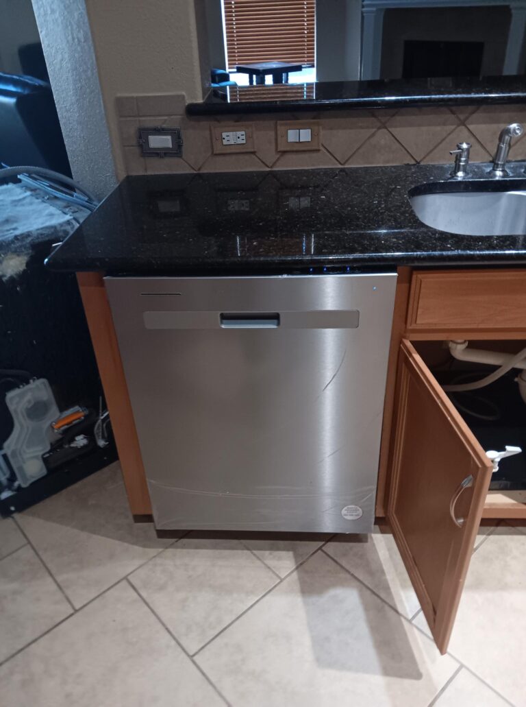 Appliance installation: A modern stainless steel dishwasher installed under a black granite countertop in a kitchen. The countertop also has a stainless steel sink with a faucet. There is a wooden cabinet with an open door next to the dishwasher, revealing the plumbing underneath the sink. Electrical outlets and a light switch are visible on the tiled backsplash above the countertop.