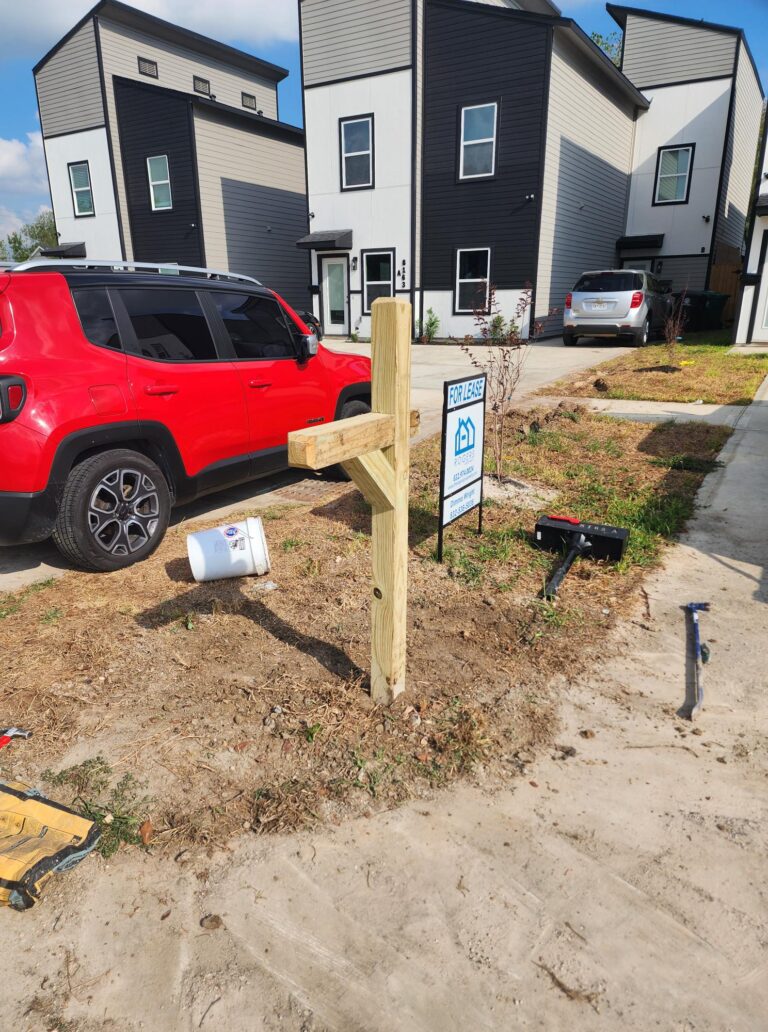 A new wooden mailbox post being installed in front of a modern townhouse. The post is securely set in the ground, with tools and materials scattered around the area, including a white bucket, a toolbox, and a wrench.