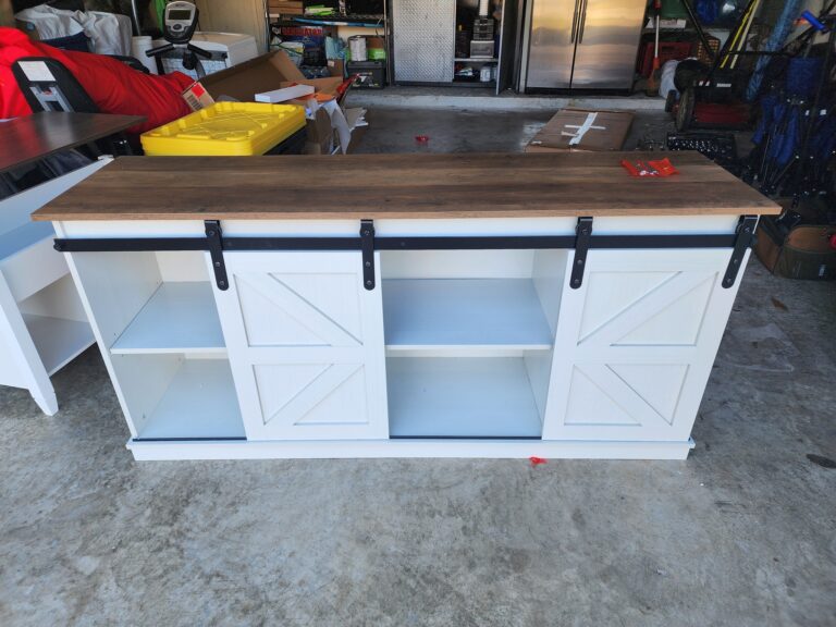 A white wooden cabinet with a dark wooden top, featuring two barn-style sliding doors on black metal tracks. The cabinet has open shelves on both sides and is situated in a garage with various tools and items in the background.