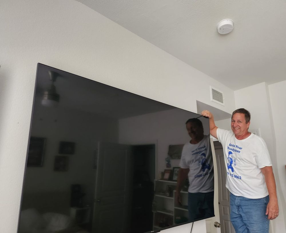 A handyman in a white T-shirt and blue jeans is standing on a step stool, finishing the installation of a large flat-screen TV mounted on a wall in a residential room. The handyman is smiling and holding the top corner of the TV. The room features white walls, a smoke detector on the ceiling, and various furnishings in the background, including a ceiling fan and framed pictures.