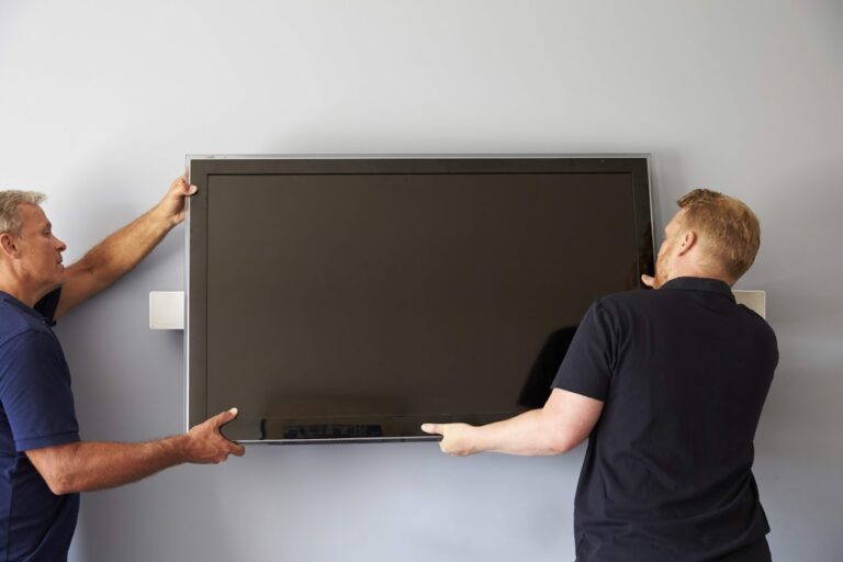 Two men are carefully mounting a large flat-screen TV on a light gray wall, ensuring it is securely positioned.